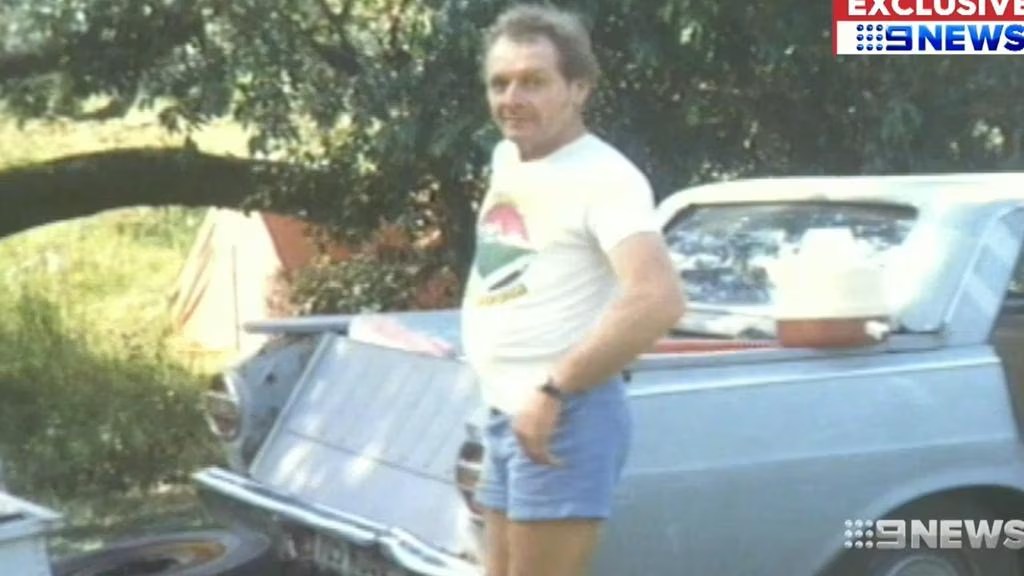 A man standing beside a vintage car in a rural setting, wearing a t-shirt and shorts.