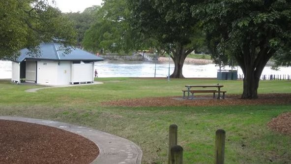 A park scene featuring a grassy area with a picnic table, a small white restroom building, and trees. In the background, there is a body of water.